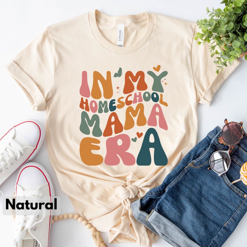 May include: A beige t-shirt with the text "In My Homeschool Mama Era" in colorful, retro-style lettering. The shirt is laid out on a white surface with a pair of white sneakers, a pair of blue denim shorts, and a wooden bead necklace.