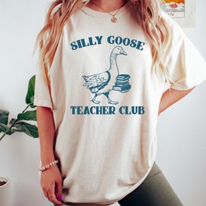 May include: A white t-shirt with a blue graphic of a goose carrying books. The text "Silly Goose Teacher Club" is printed on the shirt.