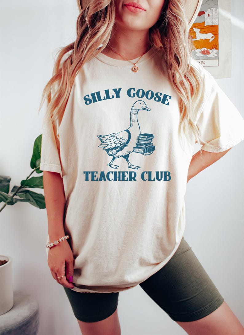 May include: A white t-shirt with a blue graphic of a goose carrying books. The text "Silly Goose Teacher Club" is printed on the shirt.