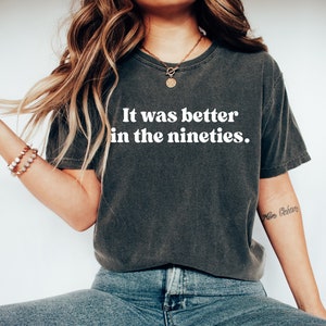 Może przedstawiać: Szary t-shirt z nadrukiem "It was better in the nineties." w kolorze białym. Koszulkę nosi osoba z długimi brązowymi włosami.