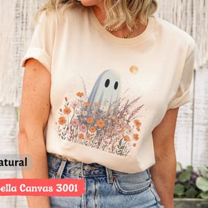 Pu&ograve; includere: T-shirt color crema con un fantasioso disegno di fantasma in mezzo a un campo di fiori selvatici e una luna piena. La maglietta ha il testo "natural" e "Bella Canvas 3001" stampato. La maglietta &egrave; in materiale morbido.