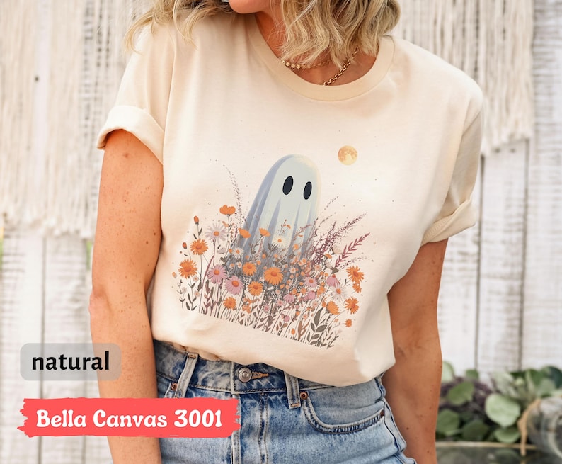 Pu&ograve; includere: T-shirt color crema con un fantasioso disegno di fantasma in mezzo a un campo di fiori selvatici e una luna piena. La maglietta ha il testo "natural" e "Bella Canvas 3001" stampato. La maglietta &egrave; in materiale morbido.