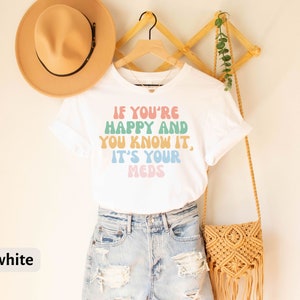 May include: White t-shirt with the text "If you're happy and you know it, it's your meds" in rainbow colors. The shirt is on a hanger with a straw hat and a woven bag.