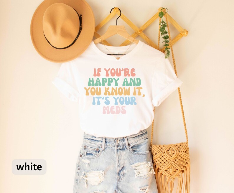 May include: White t-shirt with the text "If you're happy and you know it, it's your meds" in rainbow colors. The shirt is on a hanger with a straw hat and a woven bag.
