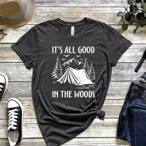 May include: A dark grey t-shirt with a white graphic of a tent in the woods. The text "It's all good in the woods" is printed on the shirt.