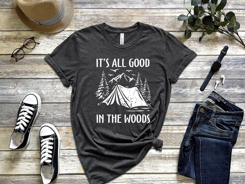 May include: A dark grey t-shirt with a white graphic of a tent in the woods. The text "It's all good in the woods" is printed on the shirt.
