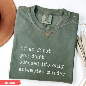 Op de afbeelding: Mosgroen Comfort Colors T-shirt met de witte tekst "if at first you don't succeed it's only attempted murder". Een gouden ketting met hanger ligt op het shirt. Maat S-P-CH.