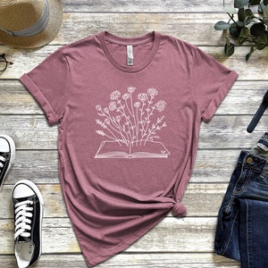 K&ouml;nnte beinhalten: Ein mauvefarbenes T-Shirt mit einer wei&szlig;en Grafik eines offenen Buches, aus dem Blumen wachsen.