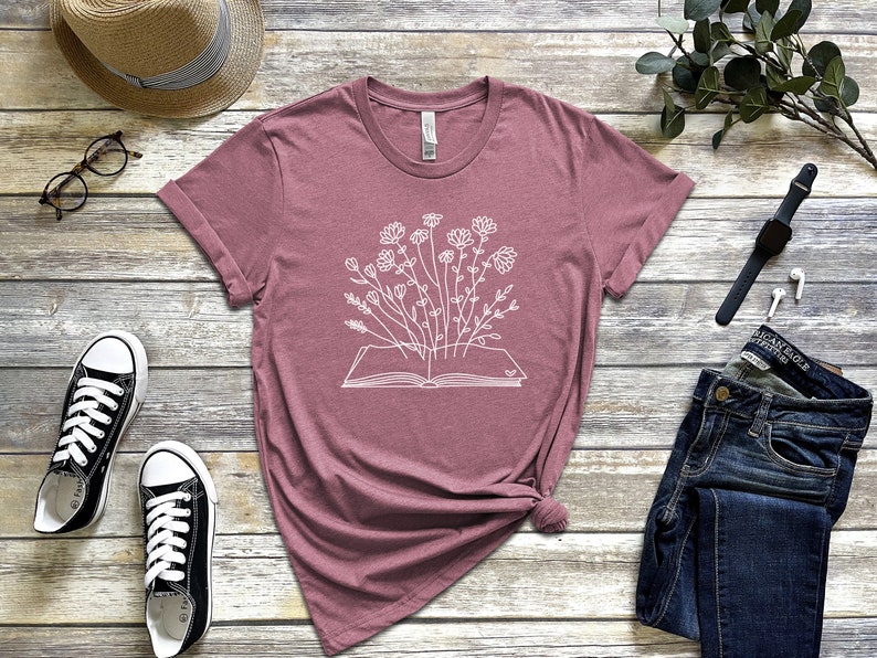 K&ouml;nnte beinhalten: Ein mauvefarbenes T-Shirt mit einer wei&szlig;en Grafik eines offenen Buches, aus dem Blumen wachsen.
