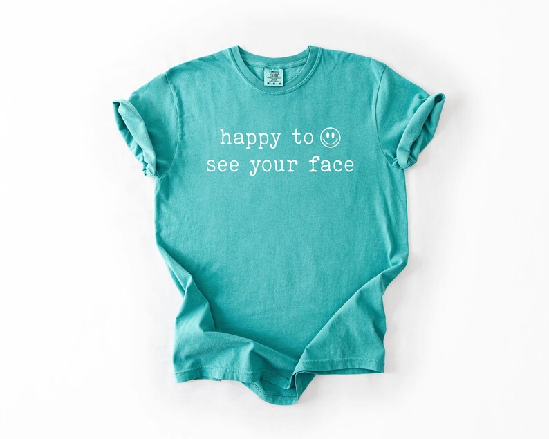 May include: A teal blue t-shirt with the text "happy to see your face" and a smiley face graphic printed in white.