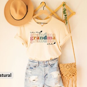 May include: A natural colored t-shirt with the text "in my grandma era" in rainbow colors. The shirt is hanging on a wooden hanger with a straw bag and a hat.