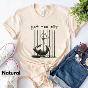 May include: A beige t-shirt with a black and white illustration of a duck in a cage. The text "got too silly" is printed above the illustration.