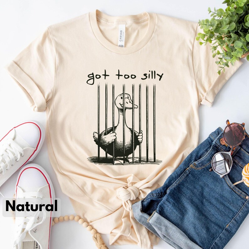 May include: A beige t-shirt with a black and white illustration of a duck in a cage. The text "got too silly" is printed above the illustration.