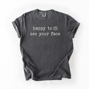 May include: A dark gray t-shirt with the text "happy to see your face" and a smiley face printed in white.
