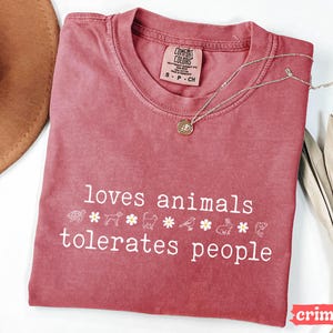 May include: A crimson red t-shirt with the text "loves animals tolerates people" in white. The shirt has a distressed look and features a row of small animal and flower icons.