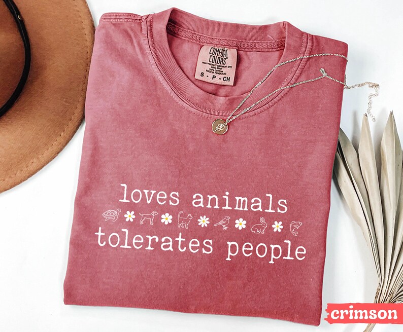 May include: A crimson red t-shirt with the text "loves animals tolerates people" in white. The shirt has a distressed look and features a row of small animal and flower icons.