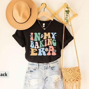 May include: Black t-shirt with the text "In My Baking Era" in colorful retro letters. The shirt is styled with a brown hat, denim shorts, and a woven bag.