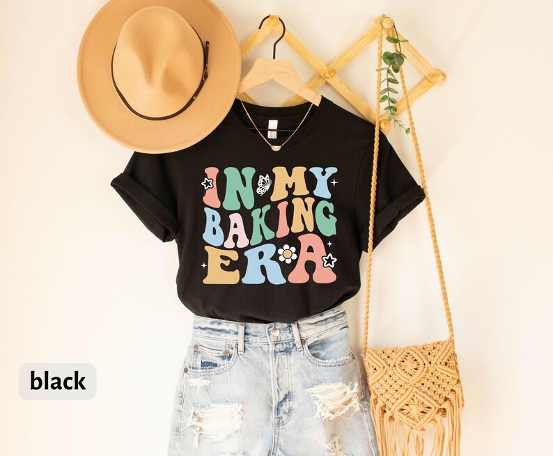 May include: Black t-shirt with the text "In My Baking Era" in colorful retro letters. The shirt is styled with a brown hat, denim shorts, and a woven bag.