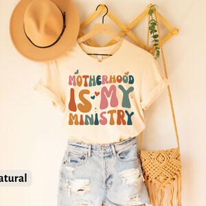 May include: A beige t-shirt with the text "MOTHERHOOD IS MY MINISTRY" in colorful, retro-style lettering. The shirt is hanging on a wooden rack with a straw bag and a hat.