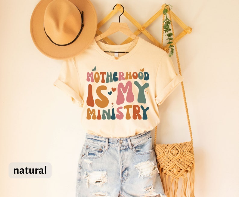 May include: A beige t-shirt with the text "MOTHERHOOD IS MY MINISTRY" in colorful, retro-style lettering. The shirt is hanging on a wooden rack with a straw bag and a hat.