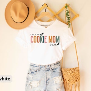 May include: White t-shirt with the text "in my cookie mom era" in colorful letters with a small heart graphic. The shirt is hanging on a wooden rack with a brown hat and a woven bag.
