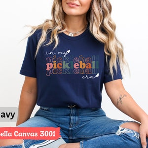 Może przedstawiać: Koszulka w kolorze granatowym z napisem "in my pickleball era" w kolorach tęczy i białymi sercami. Koszulkę nosi osoba z długimi blond włosami.