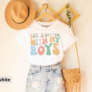 K&ouml;nnte beinhalten: Wei&szlig;es T-Shirt mit dem Text "Life is better with my boys" in bunten, Retro-Lettern.