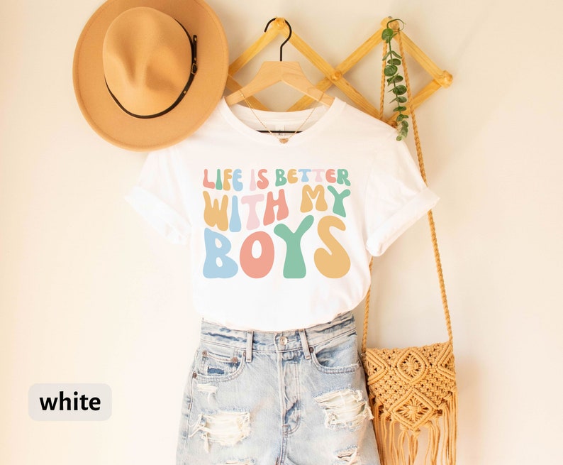 K&ouml;nnte beinhalten: Wei&szlig;es T-Shirt mit dem Text "Life is better with my boys" in bunten, Retro-Lettern.