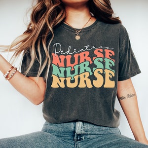 K&ouml;nnte beinhalten: Ein dunkelgraues T-Shirt mit einer Retro-Grafik, auf der "Pediatric Nurse" in gestapelten, bunten Buchstaben steht. Das Shirt wird von einer Person mit langen braunen Haaren und einer goldenen Halskette getragen.