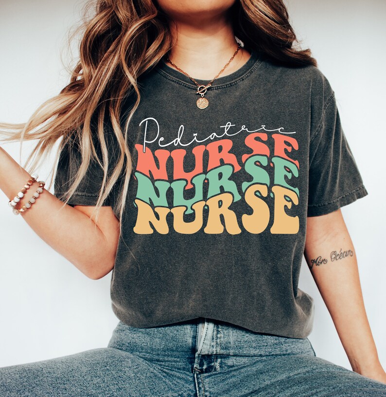 K&ouml;nnte beinhalten: Ein dunkelgraues T-Shirt mit einer Retro-Grafik, auf der "Pediatric Nurse" in gestapelten, bunten Buchstaben steht. Das Shirt wird von einer Person mit langen braunen Haaren und einer goldenen Halskette getragen.