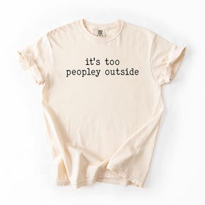 May include: A beige t-shirt with the text "it's too peopley outside" printed in black.