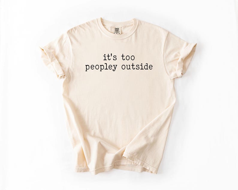May include: A beige t-shirt with the text "it's too peopley outside" printed in black.