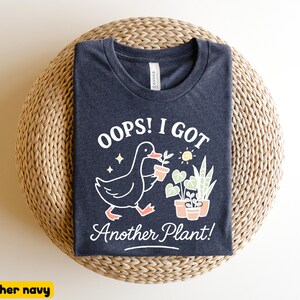 May include: Heather navy blue t-shirt with a whimsical graphic. The design features a cartoon duck carrying a potted plant, accompanied by the text "Oops! I Got Another Plant!". The shirt is laid on a woven, circular surface.