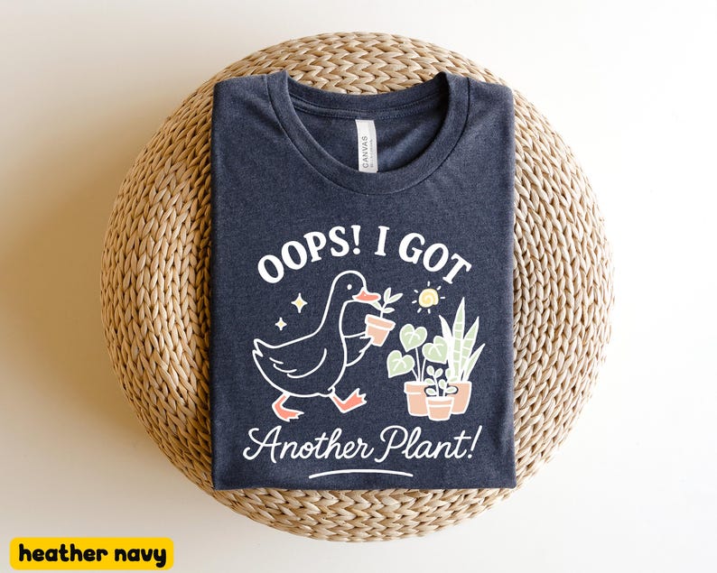 May include: Heather navy blue t-shirt with a whimsical graphic. The design features a cartoon duck carrying a potted plant, accompanied by the text "Oops! I Got Another Plant!". The shirt is laid on a woven, circular surface.