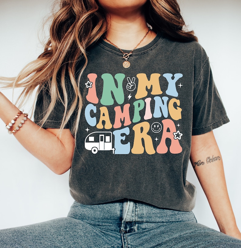 May include: A dark grey t-shirt with a retro-style graphic that says "In My Camping Era" in colorful letters. The graphic also includes a small camper van and peace sign.