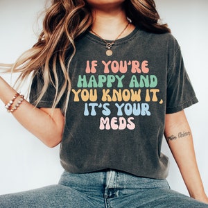 May include: A dark grey t-shirt with the text "If you're happy and you know it, it's your meds" in colorful, rounded letters.