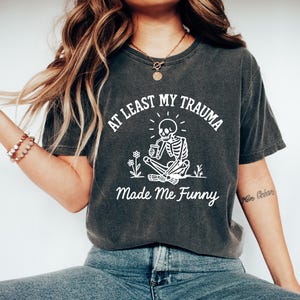 K&ouml;nnte beinhalten: Dunkelgraues T-Shirt mit dem Text "AT LEAST MY TRAUMA Made Me Funny" und einer wei&szlig;en Grafik eines Skeletts, das im Schneidersitz sitzt, eine Tasse h&auml;lt und von Blumen umgeben ist. Ein l&auml;ssiges T-Shirt.