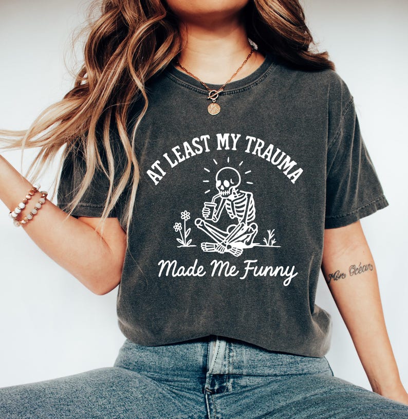 K&ouml;nnte beinhalten: Dunkelgraues T-Shirt mit dem Text "AT LEAST MY TRAUMA Made Me Funny" und einer wei&szlig;en Grafik eines Skeletts, das im Schneidersitz sitzt, eine Tasse h&auml;lt und von Blumen umgeben ist. Ein l&auml;ssiges T-Shirt.