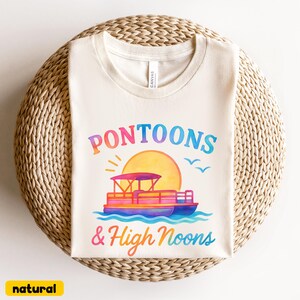 May include: Cream-colored t-shirt with the words "Pontoons & High Noons" in colorful lettering. The design includes a pontoon boat, sun, and birds. The shirt is made of a soft material and is perfect for a day on the water.