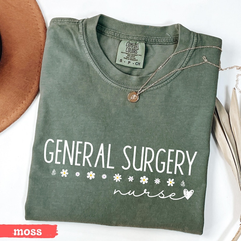 Może przedstawiać: Zielony t-shirt w kolorze mchu z napisem "GENERAL SURGERY nurse" w białym kolorze. Koszulka ma rząd białych stokrotek i małe białe serce pod napisem.