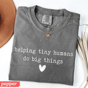 May include: A gray Comfort Colors t-shirt with the text "helping tiny humans do big things" and a heart graphic. A gold necklace with a pendant is draped across the shirt. The shirt is folded on a white surface.