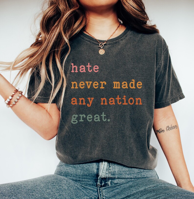 Op de afbeelding: Een donkergrijs T-shirt met de tekst "hate never made any nation great." gedrukt in kleurrijke letters. De tekst is in een vintage lettertype.
