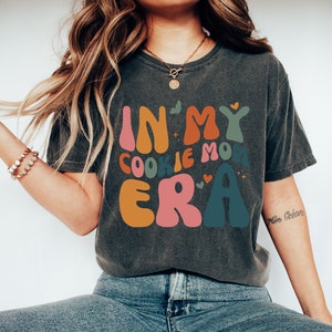 May include: A dark gray t-shirt with the text "In My Cookie Mom Era" in colorful, retro-style lettering. The shirt features a distressed look and is worn by a person with long brown hair.