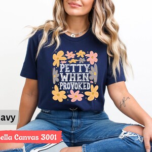 May include: Navy blue t-shirt with the text "Petty When Provoked" in a square design, surrounded by yellow, pink, and gray flowers. The shirt is a Bella Canvas 3001.