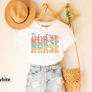 K&ouml;nnte beinhalten: Wei&szlig;es T-Shirt mit dem Text "Pediatric Nurse" in einer Retro-Schriftart mit bunten Buchstaben. Das Shirt wird von einer Person in Jeans-Shorts getragen.
