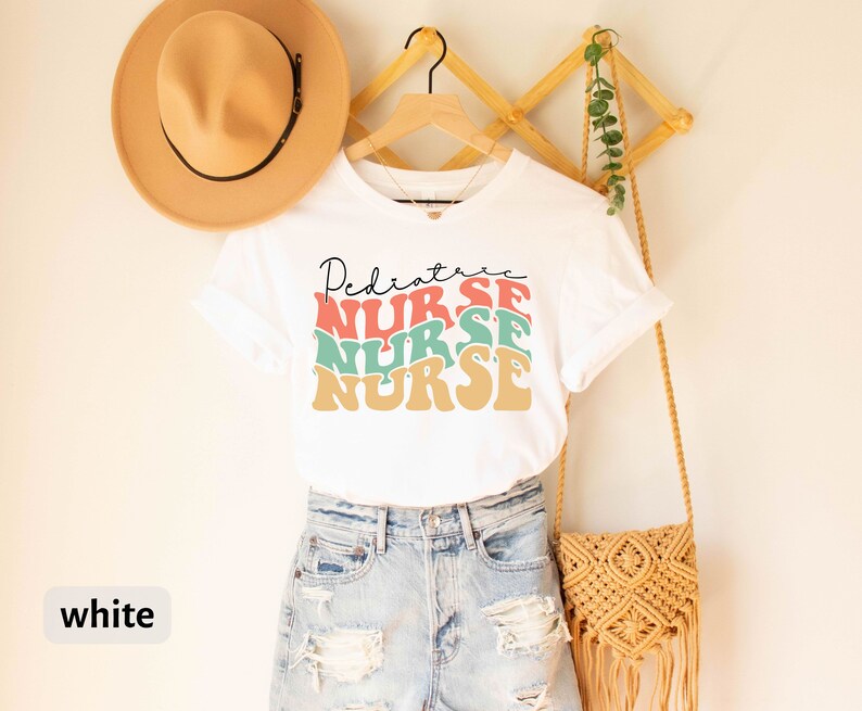 K&ouml;nnte beinhalten: Wei&szlig;es T-Shirt mit dem Text "Pediatric Nurse" in einer Retro-Schriftart mit bunten Buchstaben. Das Shirt wird von einer Person in Jeans-Shorts getragen.
