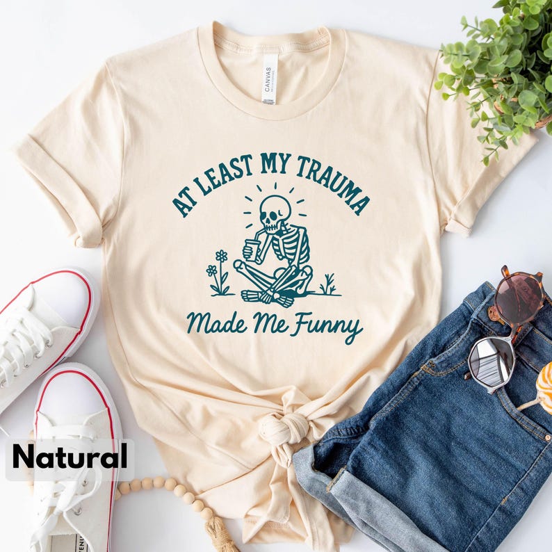 K&ouml;nnte beinhalten: Cremefarbenes T-Shirt mit dem Text "AT LEAST MY TRAUMA Made Me Funny" und einer t&uuml;rkisfarbenen Skelettgrafik. Das Shirt ist mit blauen Jeans, wei&szlig;en Turnschuhen und Sonnenbrillen versehen.