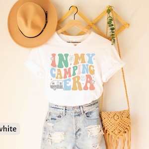 May include: White t-shirt with a colorful retro graphic that says "In My Camping Era" with a camper van, peace sign, and smiley face.