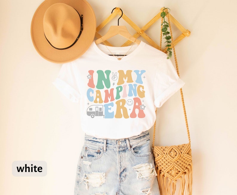 May include: White t-shirt with a colorful retro graphic that says "In My Camping Era" with a camper van, peace sign, and smiley face.