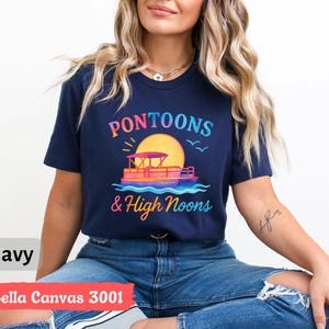 May include: Navy blue t-shirt with a colorful graphic of a pontoon boat, sun, and text that reads "Pontoons & High Noons." The shirt is labeled "Bella Canvas 3001."